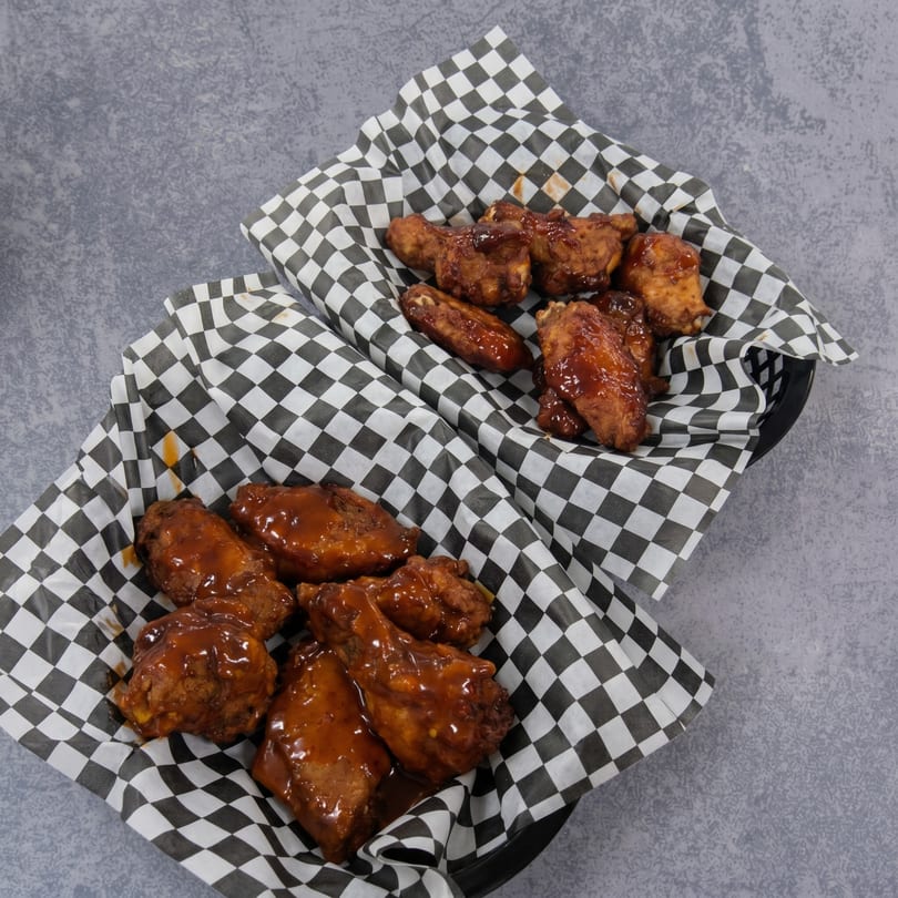Two baskets of saucy chicken wings in black and white checkered paper on a gray surface.